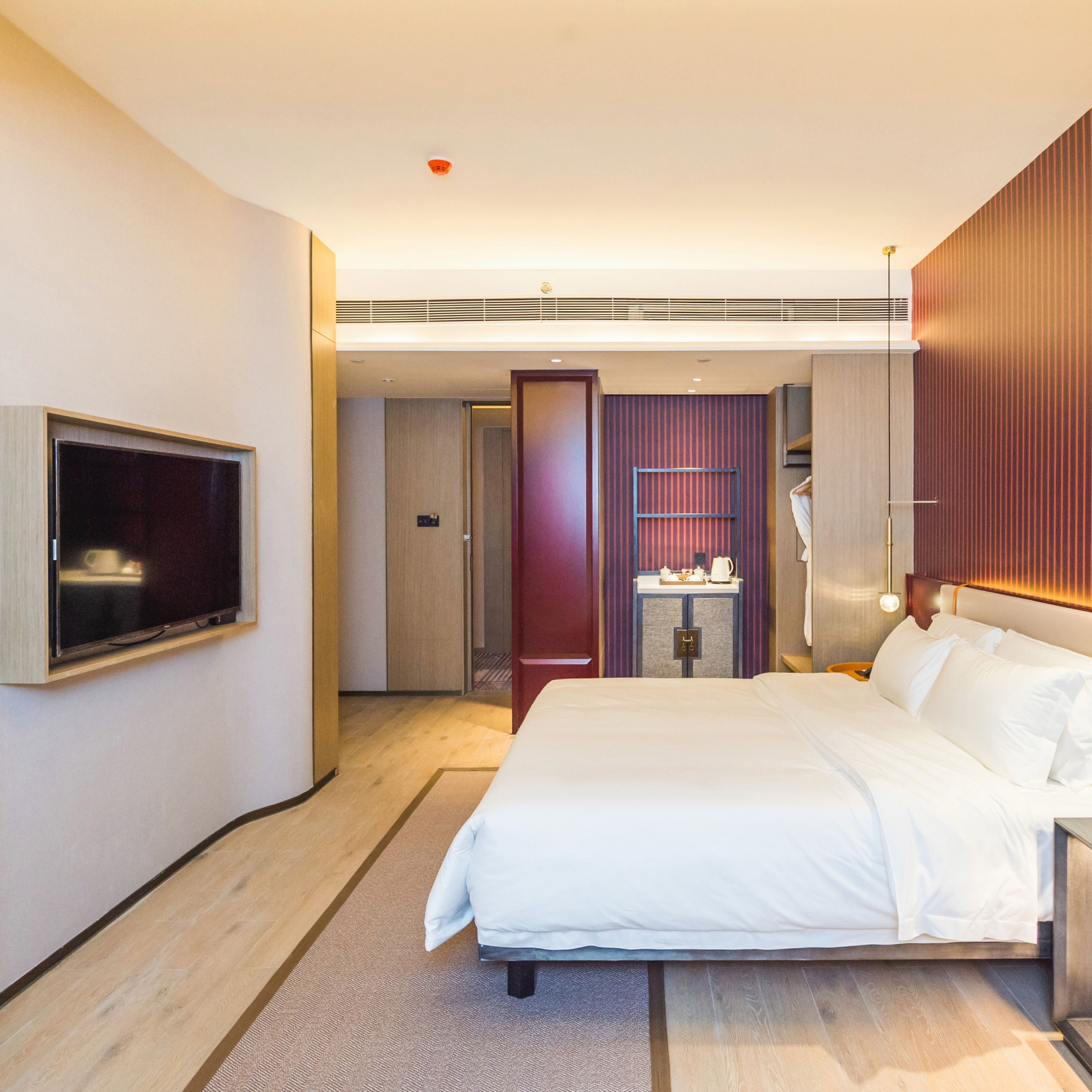 A modern hotel room with a large bed, a flat-screen TV mounted on the wall, a small table with a kettle, and a mirror above the bed.