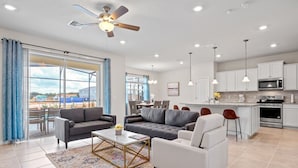 Ein geräumiges Wohnzimmer mit grauem Sofa, weißem Sessel und Couchtisch. Ein großes Fenster mit blauen Vorhängen, ein Deckenventilator und eine Küchenzeile mit weißen Schränken und einer Mikrowelle.