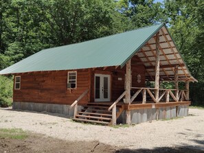 Une cabane rustique en bois, avec un toit en métal vert, une véranda dotée de balustrades en bois et un sol recouvert de gravier.