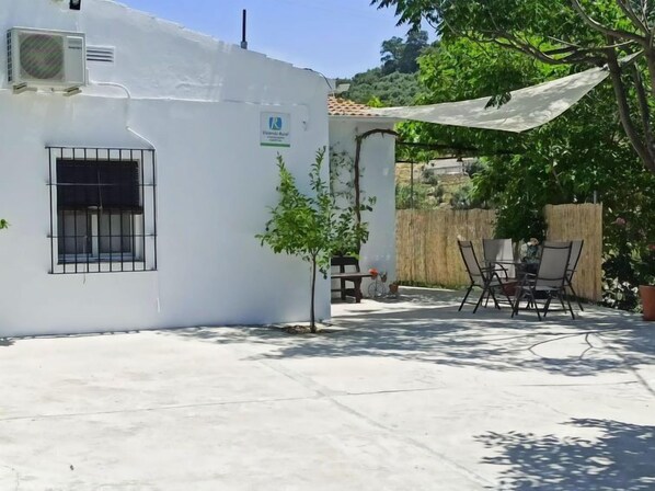A white building with a window and an air conditioning unit. There is a small tree, a table with chairs, and a wooden fence in the background.
