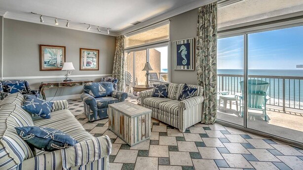 A spacious living room with striped sofas, a coffee table, and a view of the ocean through large windows. - New! Stunning Ocean View Penthouse Condo at Ocean Dunes Tower I, $300 FREE Activities Daily! (Myrtle Beach)