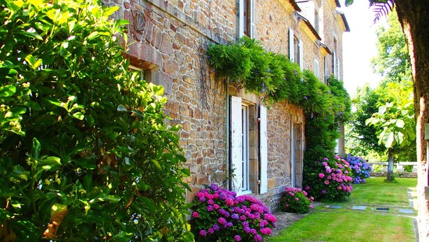 Un edificio in pietra con un vialetto in pietra che conduce all'ingresso, circondato da vegetazione lussureggiante e piante fiorite.