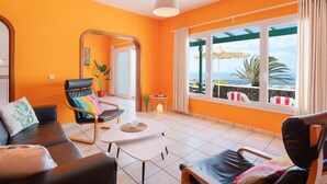 Ein Zimmer mit orangefarbenen Wänden, einem großen Fenster mit Meerblick, einem schwarzen Ledersofa, einem Holztisch und einem schwarzen Sessel mit farbigem Kissen.