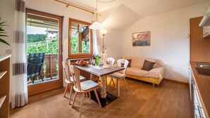 A cozy room with a dining table, chairs, a sofa, a window with a view, and a painting on the wall. - Apartment "Langeshof - Morgensonne" with Mountain View, Balcony & Garden (Kuens bei Meran, Südtirol - Italien)