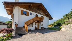 A two-story chalet with a wooden staircase, stone steps, and a mountain view. - Apartment "Langeshof - Löwenzahn" with Mountain View, Garden & Wi-Fi (Kuens bei Meran, Südtirol - Italien)