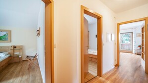 A hotel room with a wooden floor, a bed with a white mattress, a small table, and a chair. There is a door leading to another room. - Apartment "Langeshof - Löwenzahn" with Mountain View, Garden & Wi-Fi (Kuens bei Meran, Südtirol - Italien)