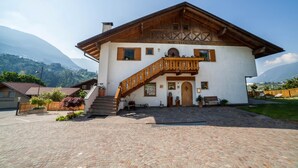 A two-story building with a wooden staircase leading to a balcony, surrounded by a paved courtyard and a mountain backdrop. - Apartment "Langeshof - Abendrot" with Mountain View, Wi-Fi, Balcony & Garden (Kuens bei Meran, Südtirol - Italien)