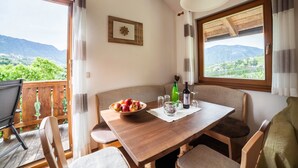 A dining area with a wooden table, chairs, a bowl of fruit, wine glasses, and a bottle. A window offers a view of greenery and mountains. - Apartment "Langeshof - Abendrot" with Mountain View, Wi-Fi, Balcony & Garden (Kuens bei Meran, Südtirol - Italien)
