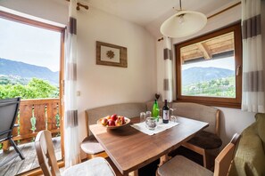 Un coin repas avec une table en bois, des chaises, un saladier de fruits, des verres à vin et une bouteille. Une fenêtre offre une vue sur la verdure et les montagnes.