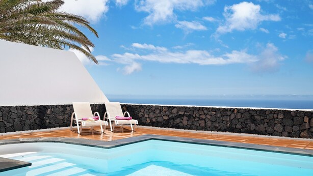 A pool with two white lounge chairs, a stone wall, and a view of the ocean. - Vacation Apartment “La Vega de Lanzarote 4” with Sea View, Pool & Wi-Fi (Tías)