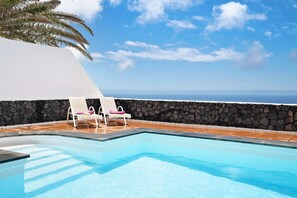 Ein Pool mit zwei weißen Liegestühlen, einer Steinmauer und Blick aufs Meer.