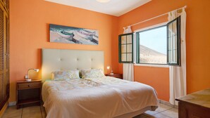 A bedroom with a bed, a nightstand, a lamp, a window with curtains, and a painting on the wall. - Vacation Apartment “La Vega de Lanzarote 4” with Sea View, Pool & Wi-Fi (Tías)