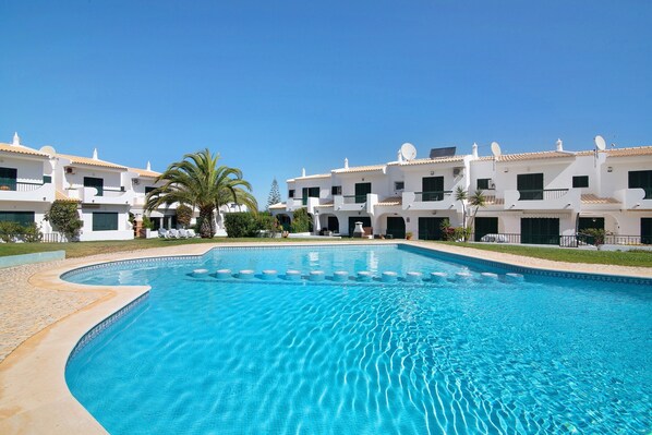 A swimming pool surrounded by white buildings with balconies, palm trees, and a clear blue sky.