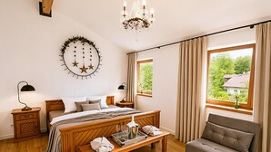 Un dormitorio con cama de madera, mesitas de noche, un banco, un sofá, una araña y vistas a zonas verdes.