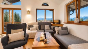 Una sala de estar acogedora con un sofá, sillones, una mesa de centro de madera y un televisor. Amplias ventanas ofrecen vista a la montaña.