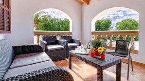 A covered outdoor seating area with wicker furniture, a table with a potted plant, and a view of greenery through arched windows. - Finca Llonguet with Mountain View, Pool & Wi-Fi (Capdepera)