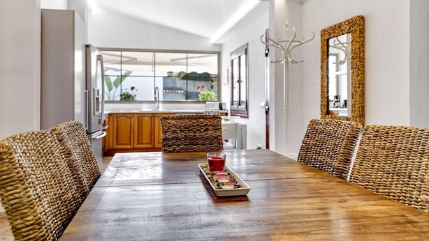 A dining area with a wooden table, woven chairs, and a view of the kitchen and outside. - Finca Llonguet with Mountain View, Pool & Wi-Fi (Capdepera)