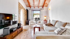 A modern living room with a TV, white sofas, a coffee table, and a stone archway leading to an outdoor area. - Finca Llonguet with Mountain View, Pool & Wi-Fi (Capdepera)