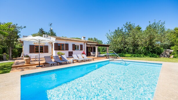 Une piscine avec des chaises longues, un bâtiment blanc aux volets bruns et un ciel bleu azur.