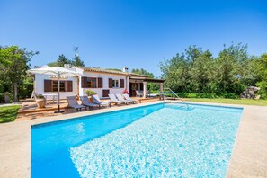 Une piscine avec des chaises longues, un bâtiment blanc aux volets bruns et un ciel bleu azur.