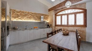 A modern kitchen with a stone backsplash, a dining table with chairs, and a large window with curtains. - Holiday Home Can Tianet with Mountain View, Pool, Wi-Fi & Terrace (Randa)