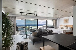 A modern hotel room with a large bed, a sofa, a dining table, and a balcony with a view of the sea.