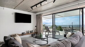Een moderne woonkamer met een grote bank, een glazen salontafel, een flatscreen televisie aan de muur en uitzicht op zee door grote ramen.