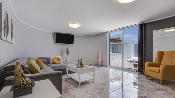A modern living room with a sofa, coffee table, and armchair, a flat-screen TV mounted on the wall, and a large sliding glass door leading to an outdoor area.