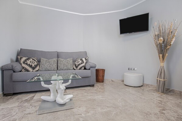 A modern living room with a grey sofa, patterned cushions, a glass coffee table, a white statue, a flat-screen TV, a decorative vase, and a wicker basket.