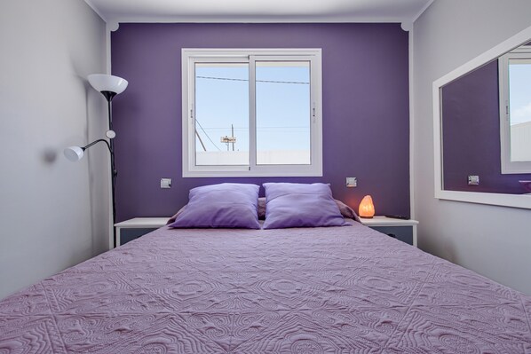Un dormitorio con cama, dos almohadas, mesita de noche con lámpara, ventana y espejo montado en la pared.