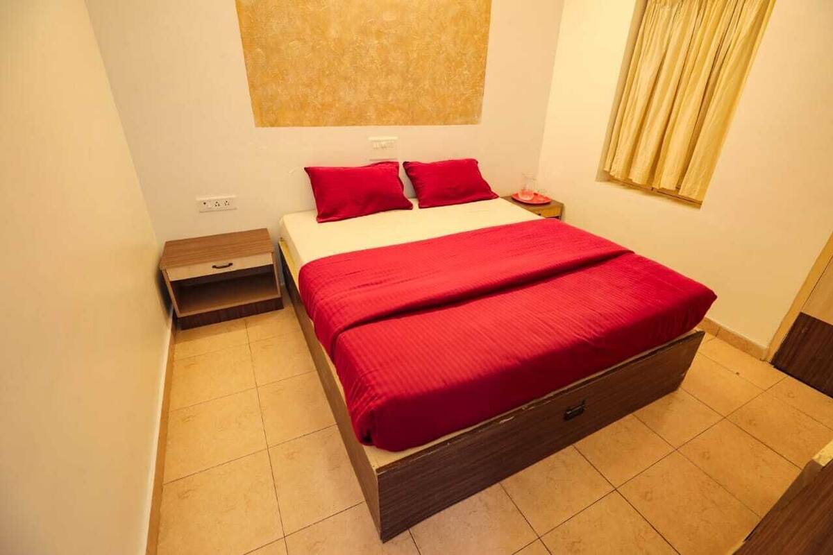 A neatly made bed with red bedding, a wooden nightstand, and a small drawer unit beside it.
