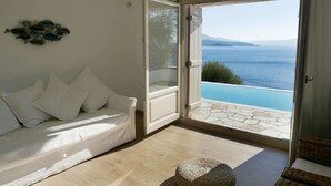 Ein Zimmer mit einem weißen Sofa, einem gewebten Hocker und einer Glastür, die zu einem Pool mit Blick auf das Meer und die Berge führt.
