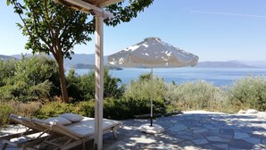 Ein sonnendurchfluteter Außenbereich mit weißem Sonnenschirm, Steinböden und Blick auf das Meer und die Berge.