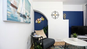 A modern living room with a blue accent wall, a round table with a plant, a grey sofa, and a painting of sailboats.