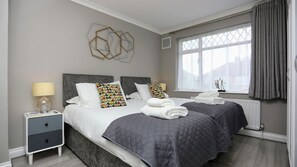 A modern bedroom with two beds, a bedside table, a window with curtains, and a geometric wall decoration.