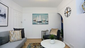 A living room with a grey sofa, a round coffee table, a painting of sailboats, a clock, and a small side table.