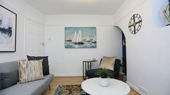 A living room with a grey sofa, a round coffee table, a painting of sailboats, a clock, and a small side table.