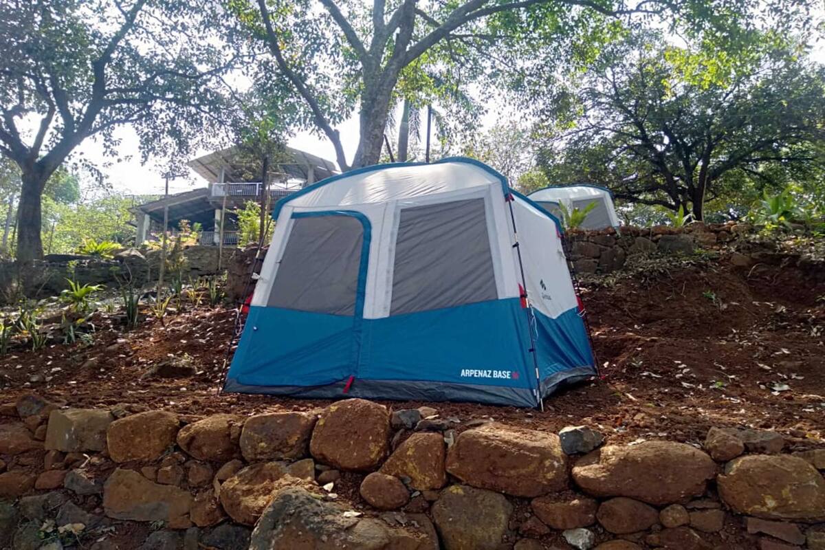 A blue and gray tent labeled "Arpenaz Base" set up on a rocky surface with trees and a building in the background.