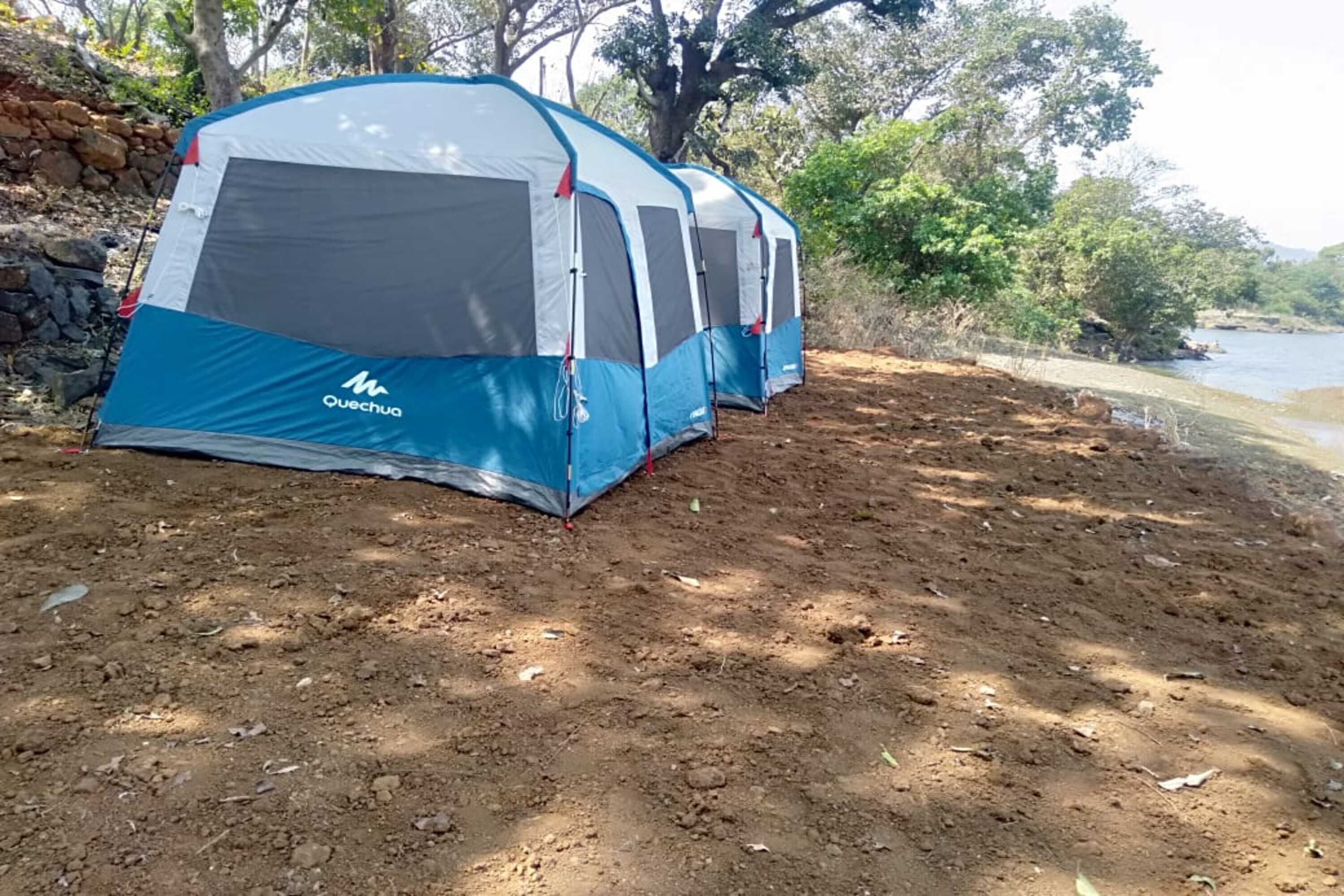 A Quechua tent set up on a dirt ground near a body of water, surrounded by trees and a stone wall.