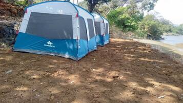 A Quechua tent set up on a dirt ground near a body of water, surrounded by trees and a stone wall.