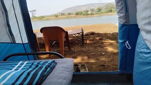 Ein Zelt mit einem Bett im Inneren, ein Stuhl davor und Blick auf ein Gewässer und Hügel.