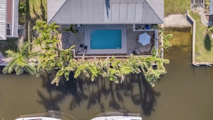 Ovenfra ses et hus med swimmingpool, omgivet af palmer og en lille bro.