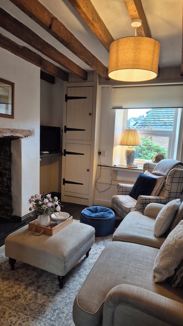 A cozy living room with a fireplace, a sofa set, a blue ottoman, and a window with a view of houses.