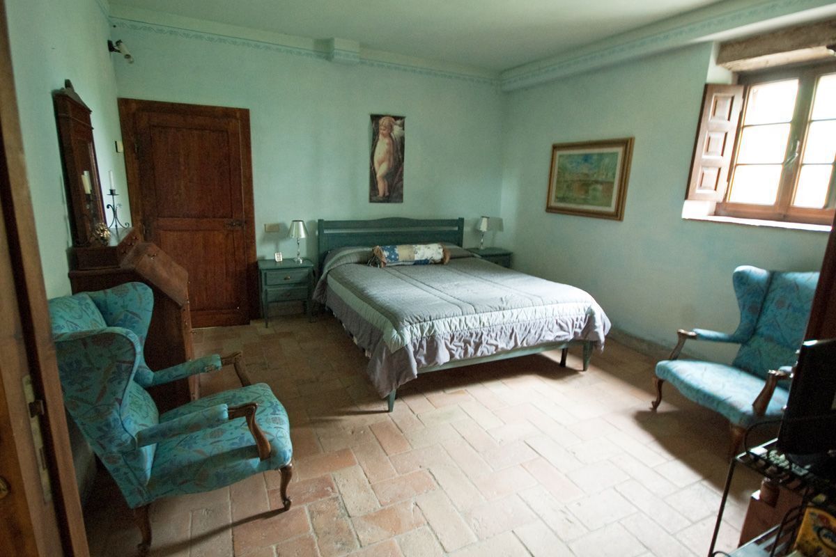 A bedroom with a bed, two chairs, a wooden wardrobe, a painting on the wall, and a window.