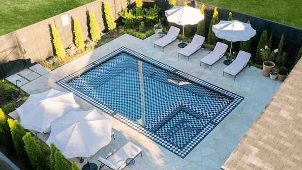 Ein rechteckiger Pool mit Schachbrettfliesen, umgeben von Liegestühlen und Sonnenschirmen.