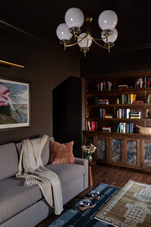 Ein Wohnzimmer mit grauem Sofa, einer Holzbücherreol, einem Chandelier und einem gerahmten Gemälde.