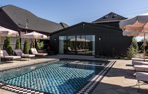 Un área de piscina moderna al aire libre con sillones de descanso, una zona de estar cubierta y un edificio con grandes ventanales.