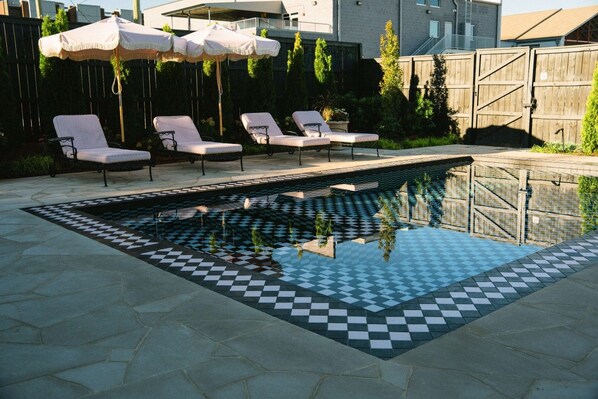 A rectangular swimming pool with a checkered tile pattern, surrounded by lounge chairs and umbrellas.