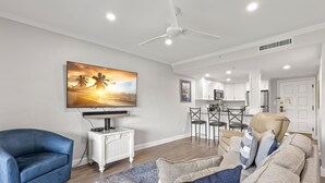 Una sala de estar con un sillón azul, un sofá beige y un televisor de pantalla plana montado en la pared.