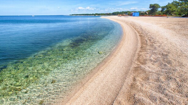 Una playa con aguas poco profundas y cristalinas, una orilla de arena y algunos edificios al fondo.